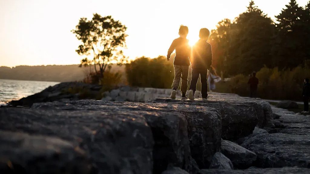 Two children walking into the sunset along lakeside rocks