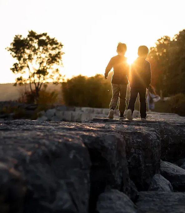 Two children walking together along lakeside rocks at sunset